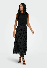 Czarny top bez rękawów z teksturowanym wykończeniem, zestawiony z czarną, długą spódnicą z kwiatowym wzorem i przezroczystą warstwą, noszony z sandałkami na paski.