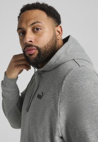 Grauer Kapuzenpullover aus weichem Material, mit einer Kängurutasche und einem schwarzen Puma-Logo auf der Brust. Nahaufnahme des Profils des Modells.