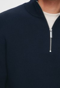 Námořnicky modrý pletený pulovr s vysokým límcem a stříbrným zipem. Tkanina má strukturovaný vzor, který zdůrazňuje teplo a pohodlí.