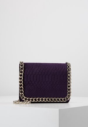 Sac à main texturé violet avec une bandoulière en chaîne argentée drapée autour de ses bords, posé sur un socle blanc sur fond uni.