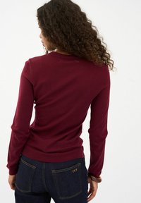 Burgundy színű, hosszú ujjú pulóver puha anyagból, kör alakú nyakkivágással, sötét farmernadrággal párosítva, arany öltésekkel és "IVY" részlettel.
