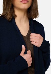 Femme ajustant un cardigan bleu marine sur un pull-over en V marron, avec une vue partielle du visage et des mains sur fond blanc.