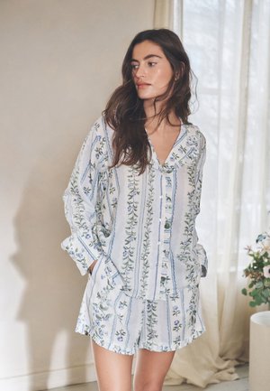 Žena s dlhými hnedými vlasmi v bielom kvetovanom pyžame s modrými a zelenými vertikálnymi vzormi stojaca v interiéri pri priehľadných závesoch a rastlinách.