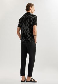 Schwarzes kurzärmliges Polohemd mit Wellenmuster, strukturierter Stoff. Kombiniert mit schwarzen, schmal geschnittenen Hosen und flachen schwarzen Sandalen. Neutraler Hintergrund.
