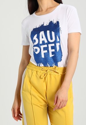 Kobieta ubrana w białą koszulkę z krótkim rękawem z niebieskim napisem graficznym oraz żółte spodnie z lnianym sznurkiem i białymi paskami po bokach.