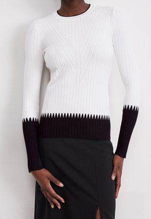 Frau trägt einen weißen, gerippten Pullover mit schwarzen Kanten an Bündchen und Saum, kombiniert mit einem schwarzen Rock mit einem vorderen Schlitz.