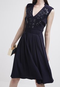 Marinblå ärmlös klänning med djup V-ringning, utsmyckad med blommig applikation vid bysten, och en flödande kjol. Håller en metallisk kuvertväska.