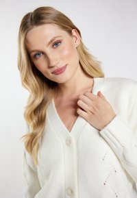 Cardigan bianco con texture, caratterizzato da scollatura a V, polsini a coste e due bottoni, con un sottile motivo a diamante lungo il corpo.