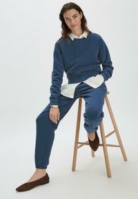 Sudadera azul con escote redondo y mangas largas, combinada con pantalones joggers azul a juego. Camisa blanca con cuello debajo y zapatos planos marrones.