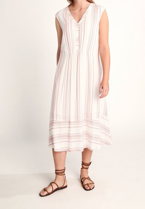 Femme portant une robe midi blanche sans manches avec des rayures verticales et horizontales roses, associée à des sandales marron en cuir avec un lien à la cheville.