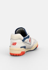 Chaussure de sport blanche avec une tige en mesh, des accents bleu marine, un logo rouge et une semelle texturée blanche et orange. Vue arrière montrant les détails du design.