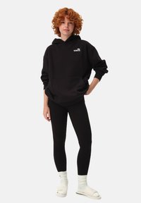 Czarna bluza z kapturem z kieszenią z przodu i białym logo, w połączeniu z czarnymi legginsami, białymi skarpetkami i beżowymi sandałami. Modelka stoi na jednolitym tle.