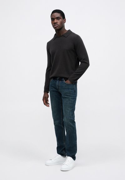 Ciemnoszara koszulka polo z długim rękawem, niebieskie dżinsy i białe trampki, stojący na tle jednolitym. Prosty, casualowy design z kołnierzykiem.