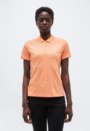 Nainen, joka pukeutunut oranssinväriseen, kuvioituun, lyhythihaiseen poolopaitaan ja mustiin housuihin, seisoo yksinkertaisen vaaleanharmaan taustan edessä.