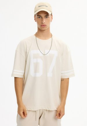 Joven con camisa beige de gran tamaño con el número 67, gorra a juego, collar de cadena y pantalones beige claro sobre fondo blanco.