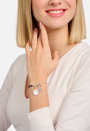 Lächelnde Frau mit blondem Haar trägt ein weißes Oberteil und ein silbernes Charms-Armband mit einem runden Blumenanhänger und Charms mit der Zahl "31".