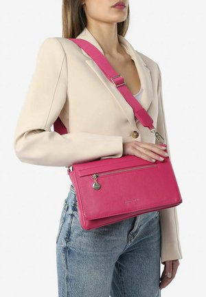 Roze getextureerde leren crossbodytas met een ritsvak aan de voorkant, verstelbare schouderband en metalen hardware. Draagt over een beige blazer.