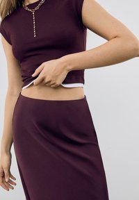 Bordeaux crop top met korte mouwen en een witte zoom, gecombineerd met een bijpassende rok. Glad textuur, minimalistisch ontwerp en subtiele gouden kettingdetail.