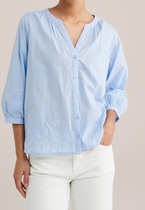 Hellblaue Bluse mit Knöpfen, dreiviertel Puffärmeln und bestickten Blumenmustern, kombiniert mit weißen Hosen. Eine Hand in der Tasche.