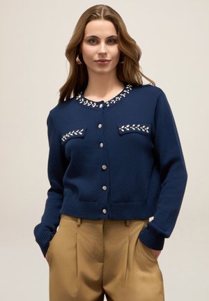 Marineblaue Strickjacke mit dekorativer Beadings um den Kragen, zwei Brusttaschen sowie gerippten Bündchen und Saum. Verschluss vorne mit Knöpfen.