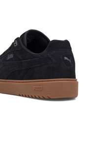 Svart mockaskor med en tan gummisula, diskret logotyp på sidan och hälen, samt en texturerad yta för ökat grepp.