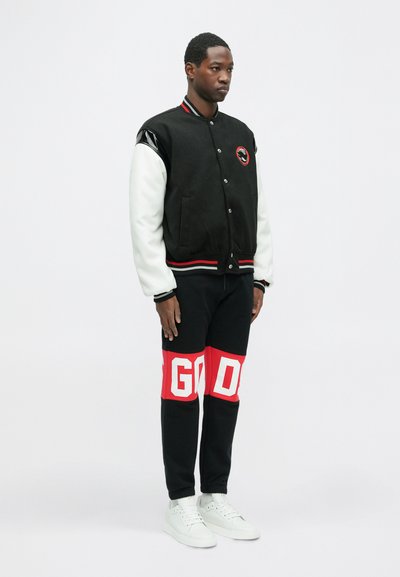 Black varsity jacket with white sleeves, red and white accents, and logo. Black pants with bold red "GOD" text. White sneakers.