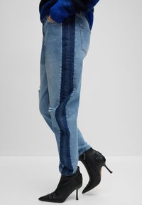 Jasnoniebieskie dżinsy z ciemnoniebieskim bocznym panelem, ze zużytymi detalami i luźnym krojem, zestawione z czarnymi, spiczastymi botkami za kostkę.