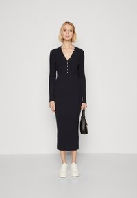 Strick-midi-Kleid in Marineblau mit langen Ärmeln, V-Ausschnitt und Knopfdetails. Trägt dazu weiße Sneakers und hält eine schwarze Handtasche.