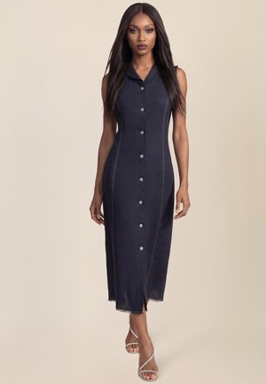 Frau mit langen, glatten Haaren, die ein ärmelloses schwarzes Midikleid mit Knöpfen und silberne, verzierte High Heels trägt, vor einem schlichten Hintergrund stehend.