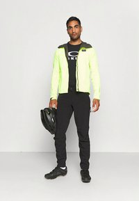 Φωσφορίζον κίτρινο τζάκετ ποδηλασίας με μαύρες λεπτομέρειες, σε συνδυασμό με μαύρο μακρύ παντελόνι και κράνος. Το τζάκετ έχει φερμουάρ μπροστά και κουκούλα.