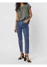 Blouse vert olive à manches courtes avec détails de plis, associée à un jean en denim bleu taille haute et des sandales noires à talons blocs.