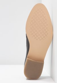 Semelle d'une chaussure habillée noire avec une semelle en caoutchouc beige présentant un motif à losanges antidérapant et un talon texturé sur fond blanc.