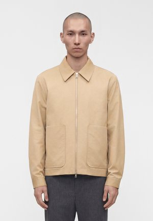 Mand med barberet hoved iført beige lynlåstrøje med store forlommer og grå bukser, står foran ensfarvet hvid baggrund.