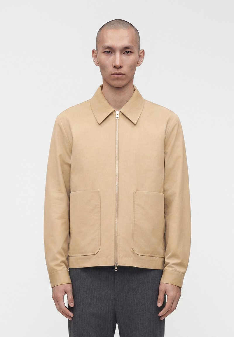 Homme au crâne rasé portant une veste beige zippée avec de grandes poches avant et un pantalon gris, debout devant un fond blanc uni.