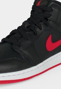 Μαύρο sneaker ψηλής κοπής με κόκκινο λογότυπο Nike, μαύρα κορδόνια και λευκή σόλα με κόκκινο πάτο, ορατό σε λευκό φόντο.