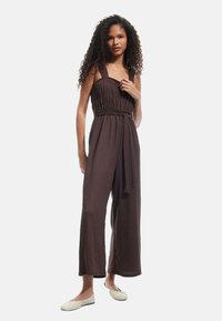 Bruine, mouwloze jumpsuit met een gerimpelde lijfje en brede pijpen, met een gestrikt riempje. De geweven textuur en de losse pasvorm vergroten het comfort.