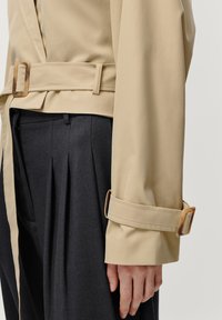 Beige trenchcoat met riem, grote manchetten en schildpadgesp, gecombineerd met donkere geplooide broeken, die een zachte textuur en op maat gemaakt ontwerp tonen.
