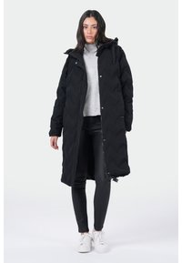 Zwarte, lange pufferjas met een gewatteerd design, capuchon en rits. Draagt over een grijze trui en zwarte broek, gecombineerd met witte sneakers.