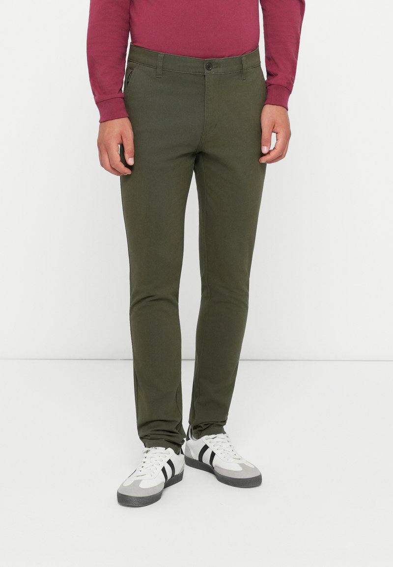 Oliva zöld, slim-fit chino nadrág sima textúrával, elülső zsebekkel, övhurkokkal és egygombos záródással. Fehér sportcipővel viselve.