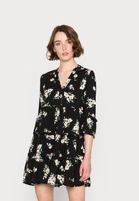 Vestido negro com flores, decote em V, mangas três quartos, volantes em camadas e um padrão de flores cor-de-rosa e brancas, feito de tecido leve.