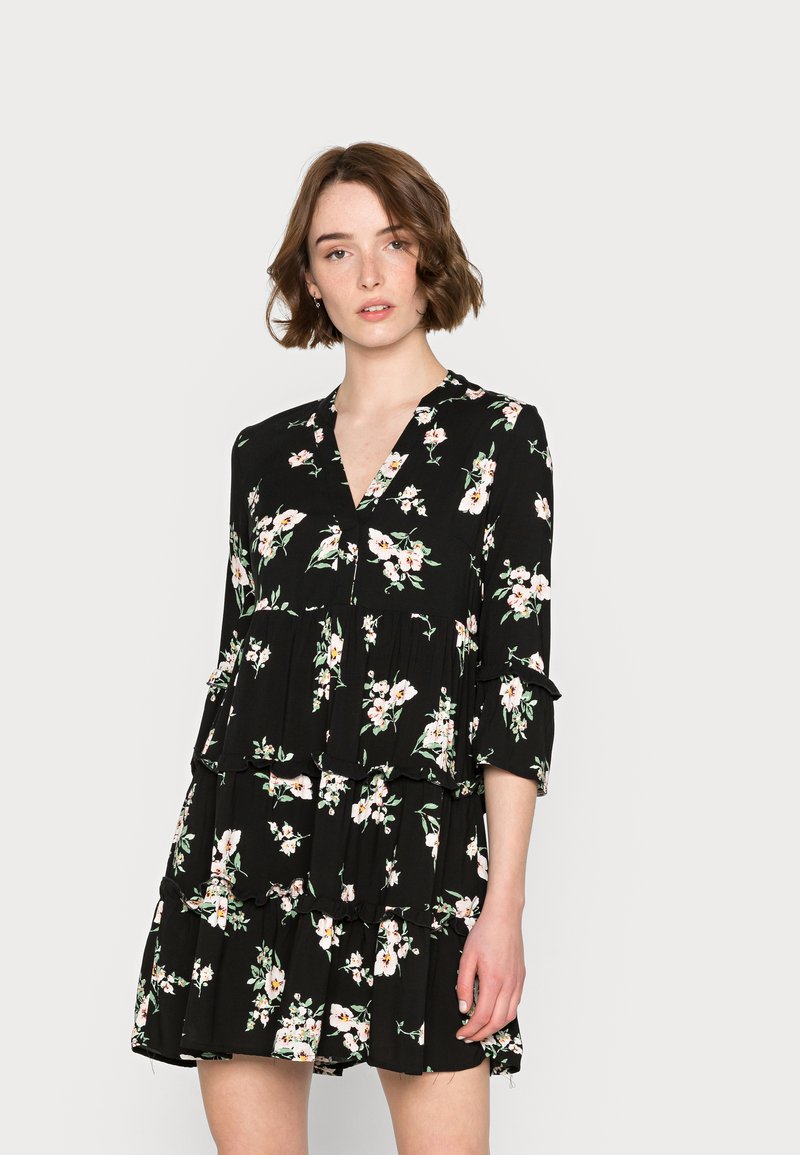 Vestido negro com flores, decote em V, mangas três quartos, volantes em camadas e um padrão de flores cor-de-rosa e brancas, feito de tecido leve.