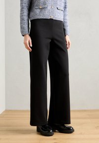 Pantalons noirs à larges jambes en tissu doux avec une texture lisse. Présentant une taille haute et associés à des chaussures noires massives.