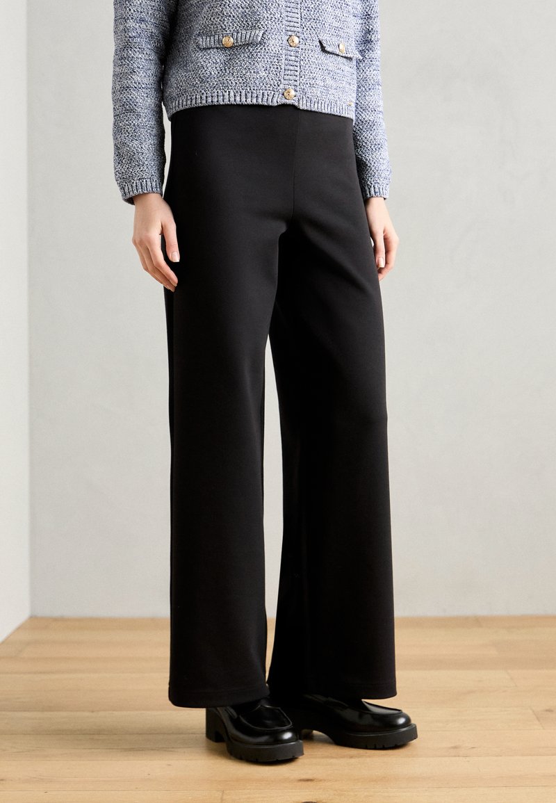 Pantalons noirs à larges jambes en tissu doux avec une texture lisse. Présentant une taille haute et associés à des chaussures noires massives.