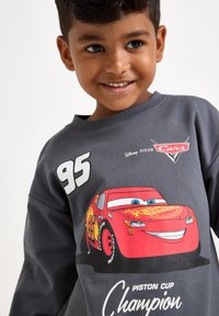 Harmaa huppari, jossa on värikäs "Cars"-design, iso Lightning McQueen -grafiikka, numero 95 ja teksti "Piston Cup Champion".