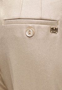 Μπεζ ύφασμα με διακριτική λάμψη, που διαθέτει τσέπη με καπάκι και διακοσμητικό κουμπί με λεπτομερή σχέδιο, μαζί με μεταλλικό διακοσμητικό στοιχείο σε σχήμα "M".