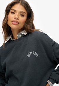 Donkerblauwe sweatshirt met een 'SUPERDRY' logo in lichtgrijs. Heeft een ronde hals en is gelaagd over een blauw-wit gestreept overhemd met kraag.