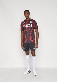 Červený a čierny futbalový dres s logom "INEOS", kombinovaný s čiernymi šortkami a bielymi ponožkami. Nosí sa s bielymi futbalovými topánkami.