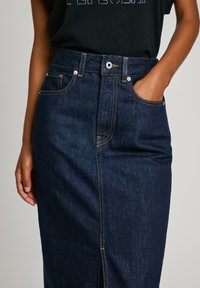 Gonna midi in denim blu scuro con cuciture visibili, spacchi frontali e tasche anteriori. Design a vita alta con chiusura a bottone e zip.