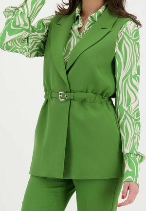 Mujer con un chaleco verde brillante con cinturón sobre una camisa de manga larga verde y blanca con estampado, con pantalones verdes a juego.