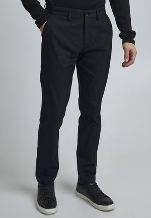 Black trousers made of cotton blend fabric, featuring a straight-leg design, front pockets, and a button closure. Paired with black sneakers.
